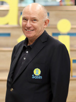 Ian Fenwick of Sasin School of Management, an older gentleman with a light complexion, friendly smile, and blue eyes, and a clean-shaven head with slight gray hair at temples. He wears a black suit jacket with Sasin's yellow and blue logo over a black polo shirt. He stands in front of a wide set of light-colored wooden steps with the yellow letters "Sasin" descending diagonally with one letter on each step.