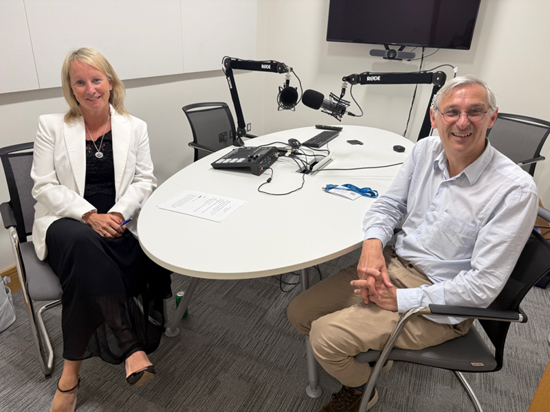 Eileen McAuliffe (left) and Andrew Jack (right) sitting at a table with audio recording equipment, smiling at camera