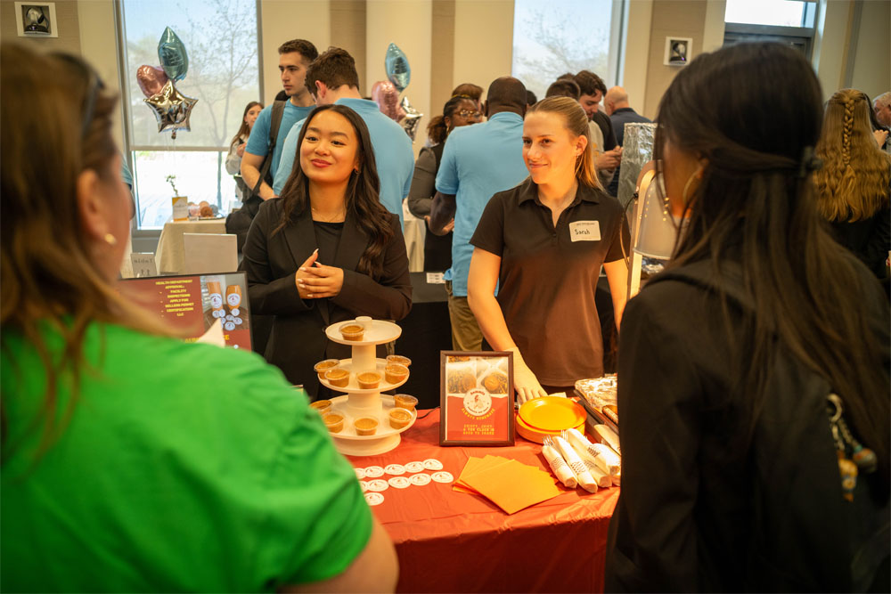 Two young women stand behind a red-covered table at the Rohrer New Venture & Entrepreneurship Expo, talking with attendees whose backs are turned to the camera about their product Sammi's Sandwich and Sauce. The table holds a multitiered stand with small sample cups of sauce, plates and napkins, and a display board, with people and balloons in the background.