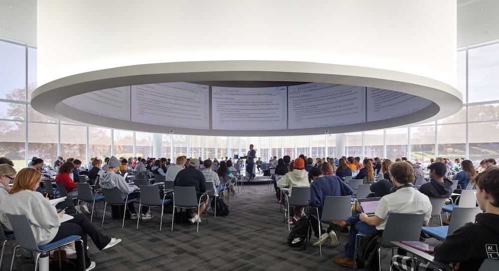 Wide shot of a full classroom in a big room with glass walls. Overhead is a large cylindrical feature with monitors built inside.