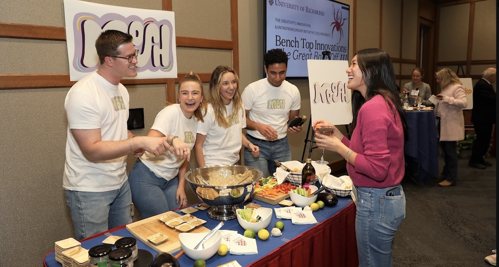 Smiling students in white shirts share baba ghanoush samples with visitors at a food fair.
