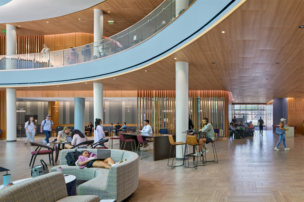 The bright, open atrium of the Owen School's new building includes a sweeping, curved upper balcony with a glass railing and wood-paneled underside. On the ground floor, students study, chat, and walk across a wood-look tile floor and sit on curved, gray upholstered sectional sofa and bench in the foreground and long work tables with modern barstools and informal seating clusters in the background in front of a slatted wood accent wall.