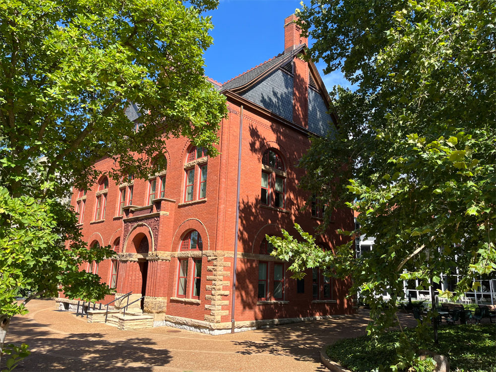 A low-angle, exterior shot shows Old Mechanical, the 1880s historic three-story, red-brick building on Vanderbilt's campus and part of the Owen School of Management's facilities. The building's architecture is highlighted by a light-colored stone foundation, tall arched windows with red trim, and a steep gabled roofline with slate-colored shingles. It is surrounded by a paved walkway in the foreground, as well as leafy trees whose shadows dapple the brick walls.